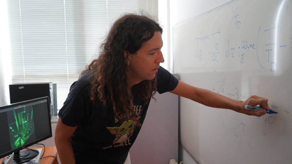 Foto de Santiago Oviedo en su despacho, junto a una imagen del experimento que ha validado su investigación teórica, cuya fórmula principal aparece en la pizarra.